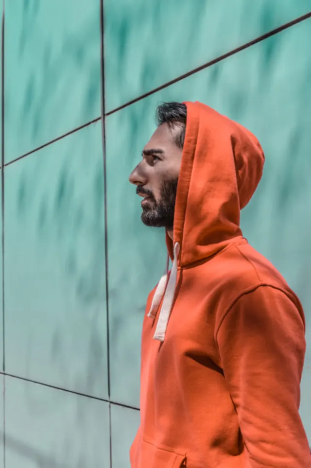 Man in an orange hoodie standing against a teal wall