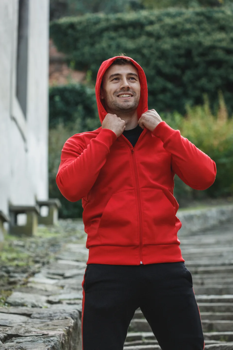 Smiling man pulling up the hood of a red zip-up jacket on stone steps